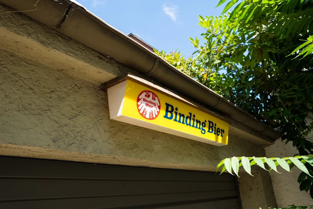 Leuchtschild mit dem Schriftzug 'Binding Bier' und rotem Logo an einer Hauswand unter einem Dachvorsprung mit grünen Blättern.