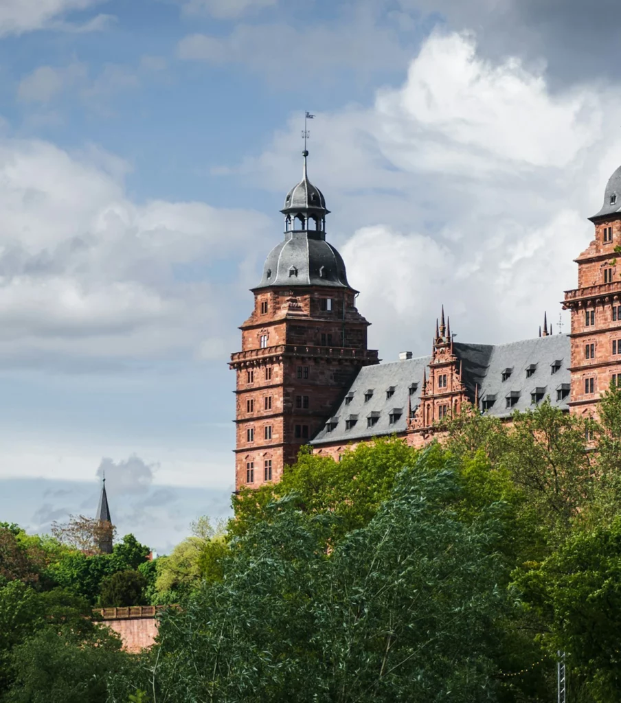Das Schloss Johannisburg in Aschaffenburg mit seinem markanten Turm und dem steinernen Dach, umgeben von grünen Bäumen unter einem bewölkten Himmel